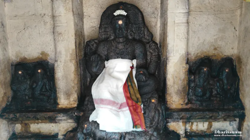 Black stone seated idol with a white cloth, flanked by small carved figures on both sides