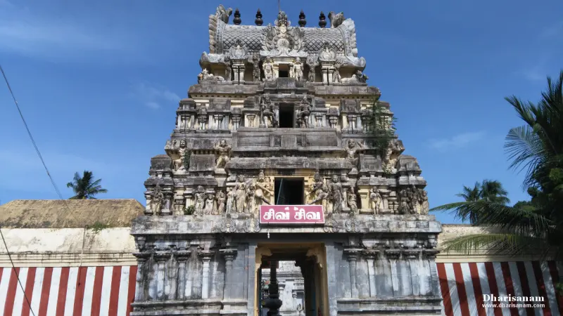 Three-tiered gopuram with carved figures against a blue sky, with a red and white striped wall on either side