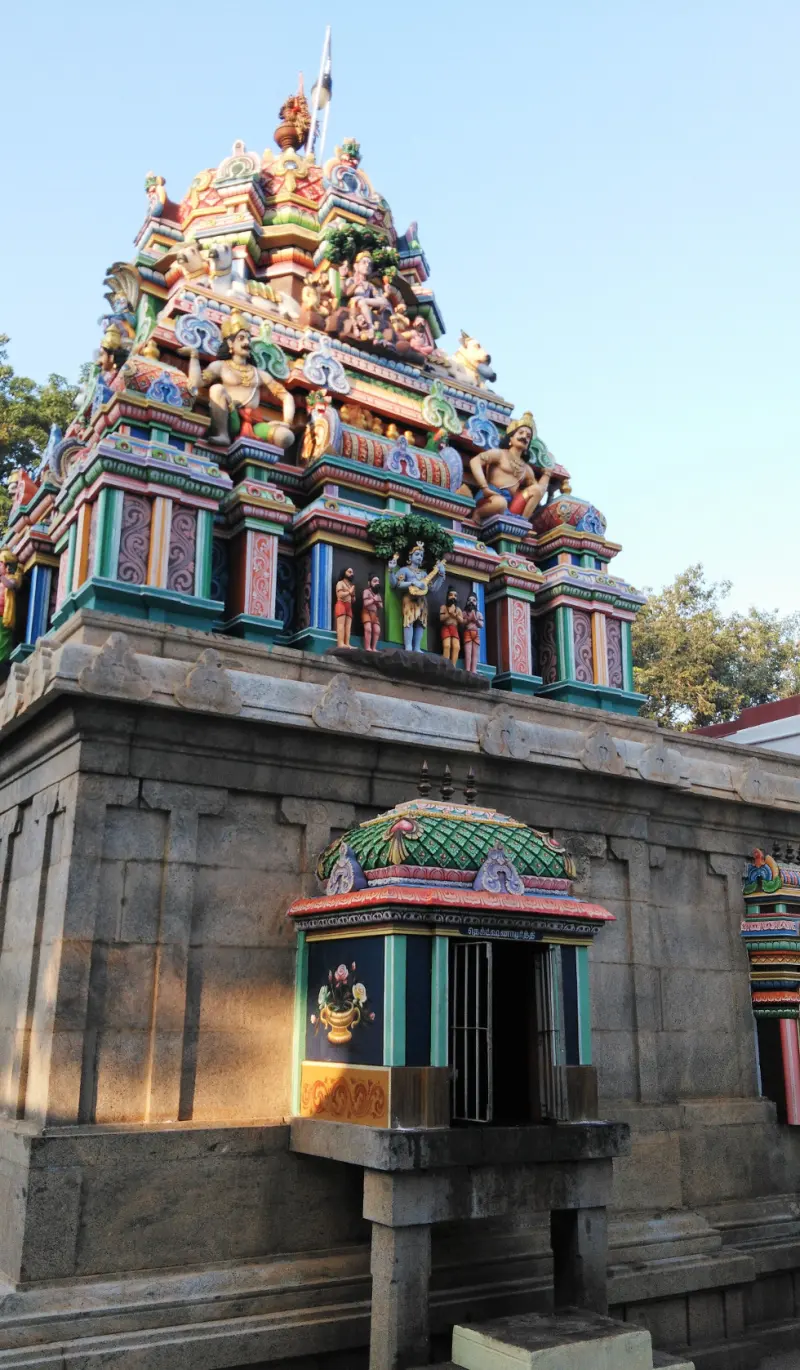 A colourful tower covered with sculpted figures above a stone wall, with a small shrine on the wall.
