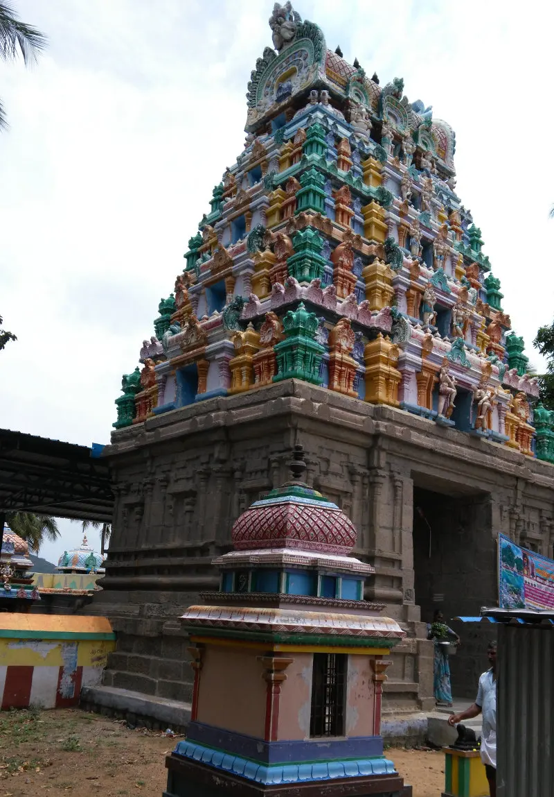 A painted gopuram over a stone base, a small domed shrine in front and a few people near the entrance.