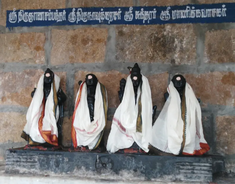 Four dark figures in white cloth stand in a row beneath a blue name board in Tamil