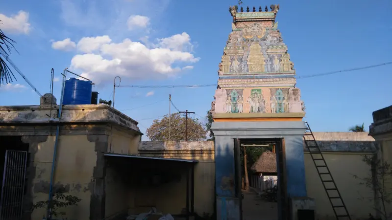 A painted tower with sculptures rises above a gateway in a yellow compound wall; a ladder leans on the wall.