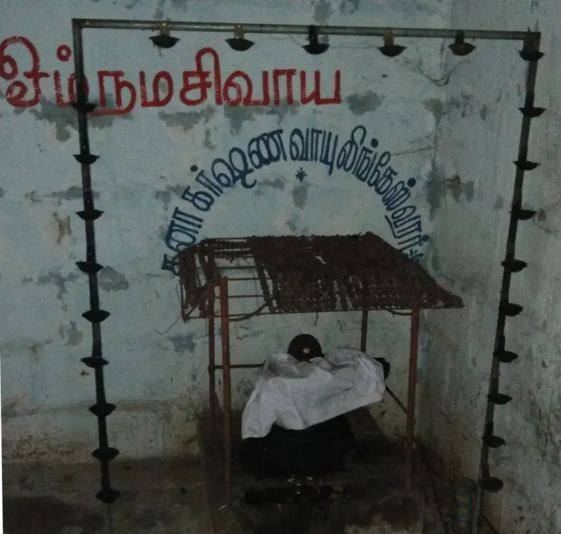 A lingam wrapped in white cloth under an iron frame with lamps, with Tamil lettering on the wall behind.