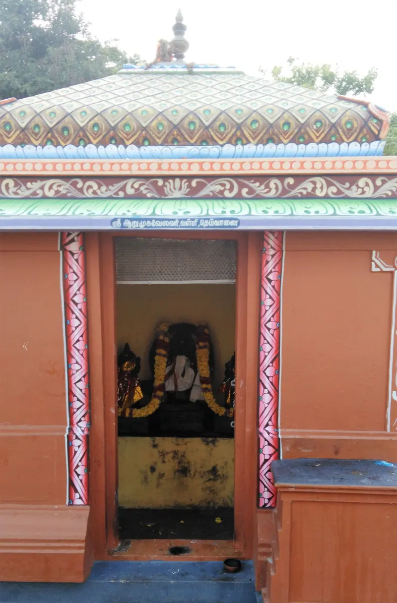 A small orange shrine with garlanded idols inside; the sign above names Arumugavelavar, Valli and Deivanai.