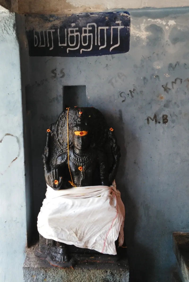 Black Veerabadrar idol with a kumkum mark, in white cloth, below a Tamil name board