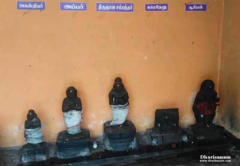 Five small black idols in a row before an orange wall with a name board above each, one dressed in red