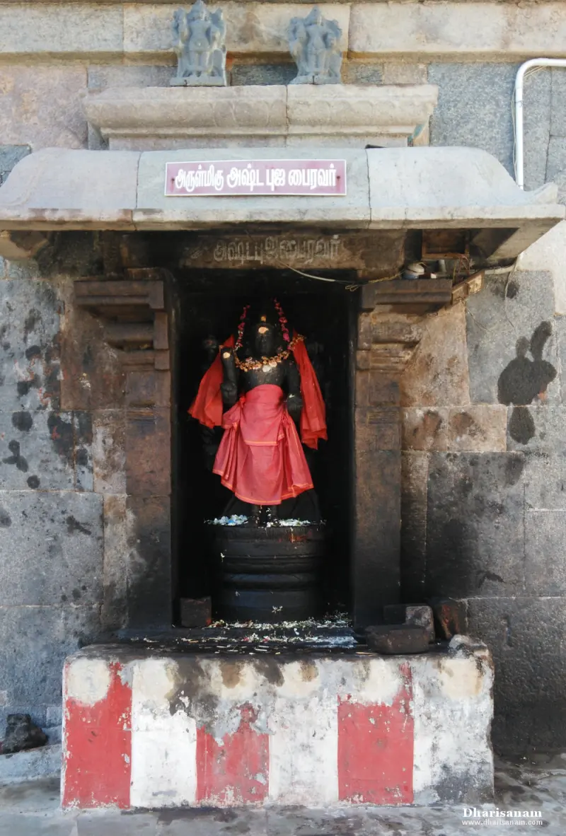 A dark idol in a red cloth standing in a stone niche with a name board above, on a red and white base.