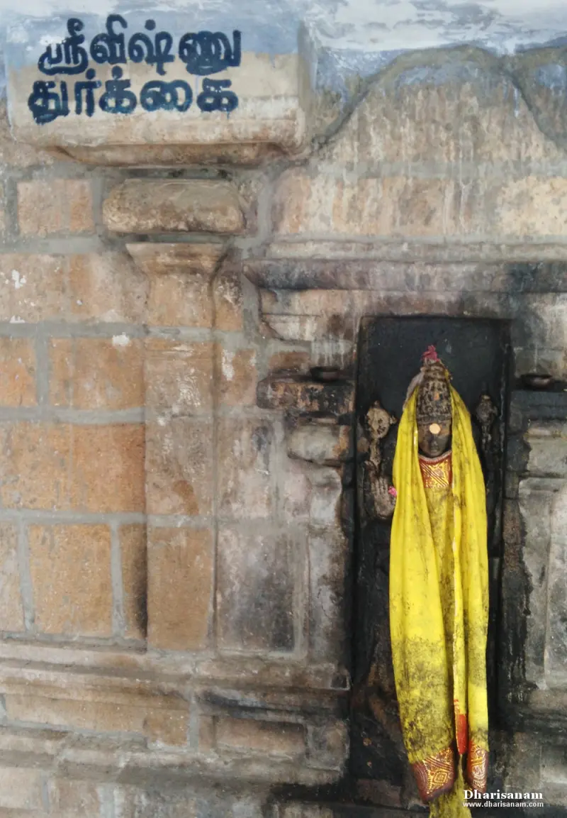 A small idol in a yellow cloth in a niche of a granite wall, with a Tamil name board above it