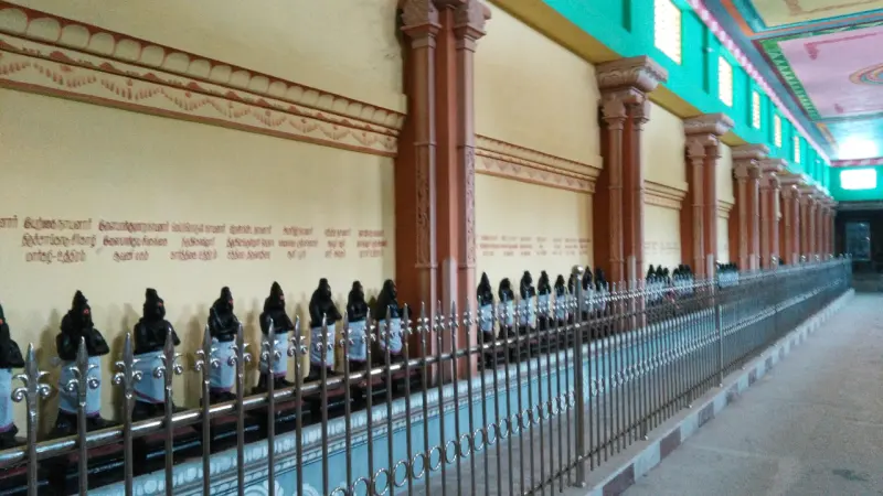 A long row of small dark idols behind a steel railing along a yellow corridor wall with painted text.