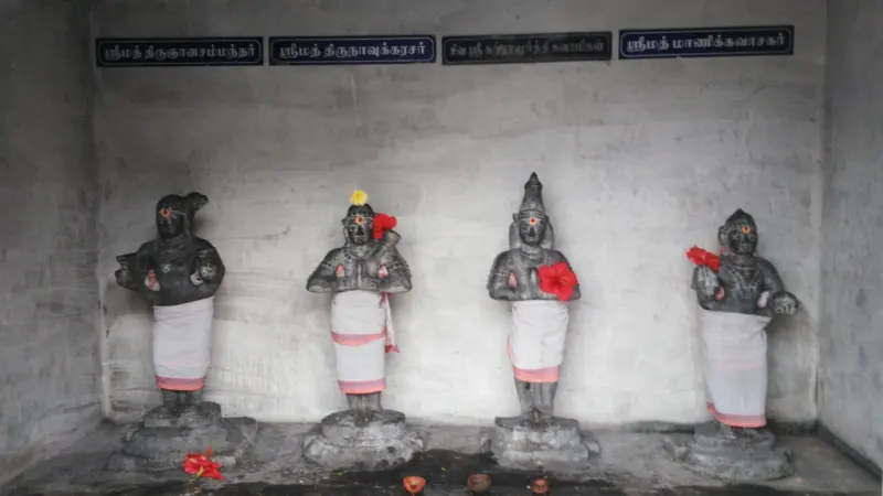 Four dark stone figures in white cloth stand on bases in a plain room, with Tamil name boards above each.