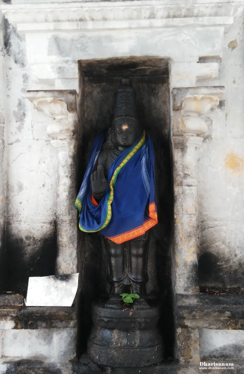 Dark standing idol in a blue and orange cloth, set in a white stone niche with pillars at its sides