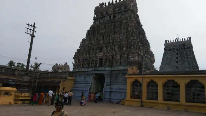 A grey stone gopuram with a smaller tower behind, seen from a courtyard with devotees.