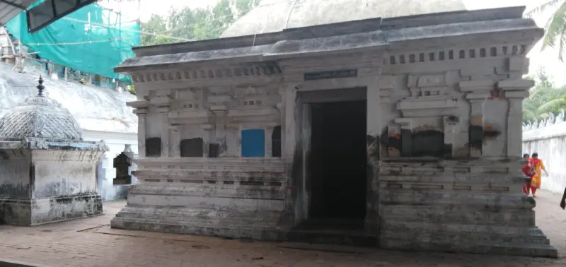 A low stone shrine with a dark open doorway and two people walking beside it.