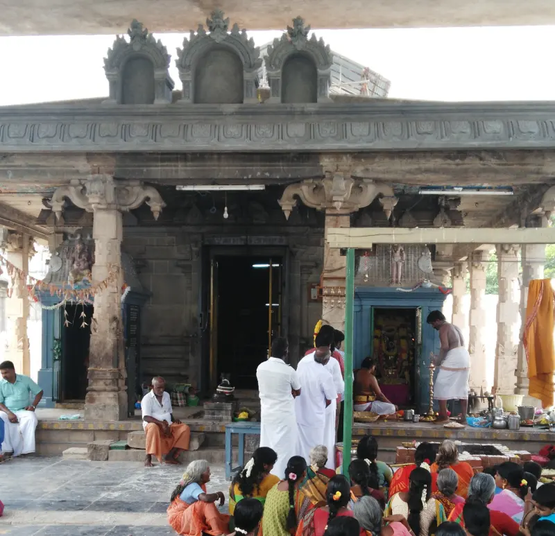 Devotees seated on the floor before a pillared mandapam while a priest conducts a pooja at a side shrine