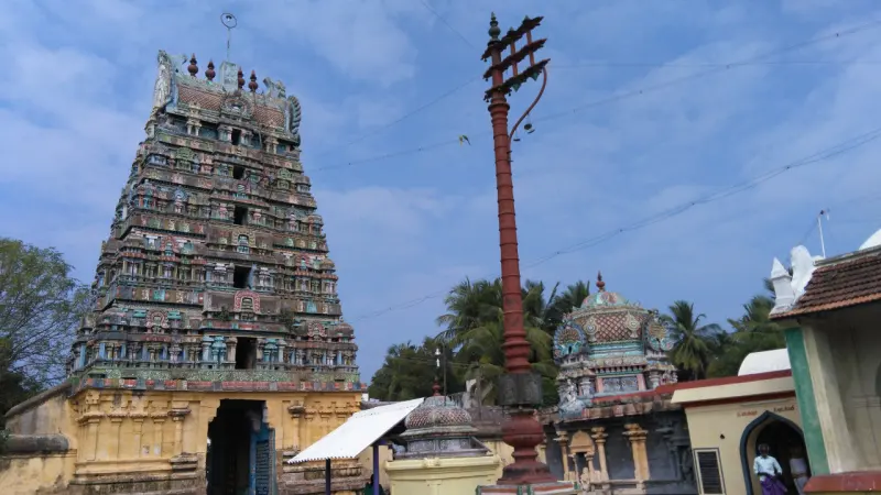 A tall painted tower rises above a yellow gateway beside a red flagstaff, with a small domed shrine at right.