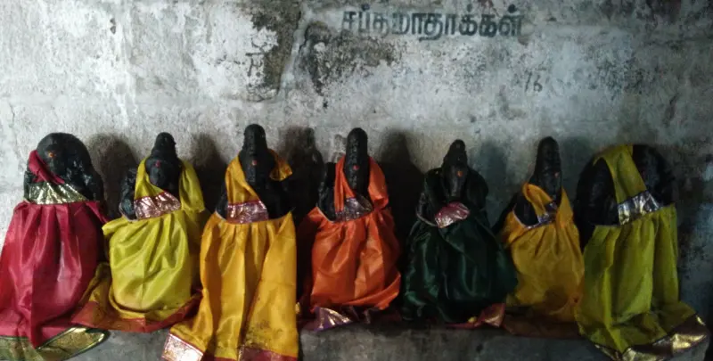 Seven dark stone idols in a row, dressed in red, yellow, orange and green cloth, with a Tamil label above