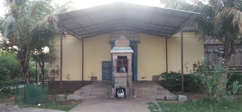 A small stone shrine with a coloured top stands under a tin roof in front of a yellow wall, among coconut palms.