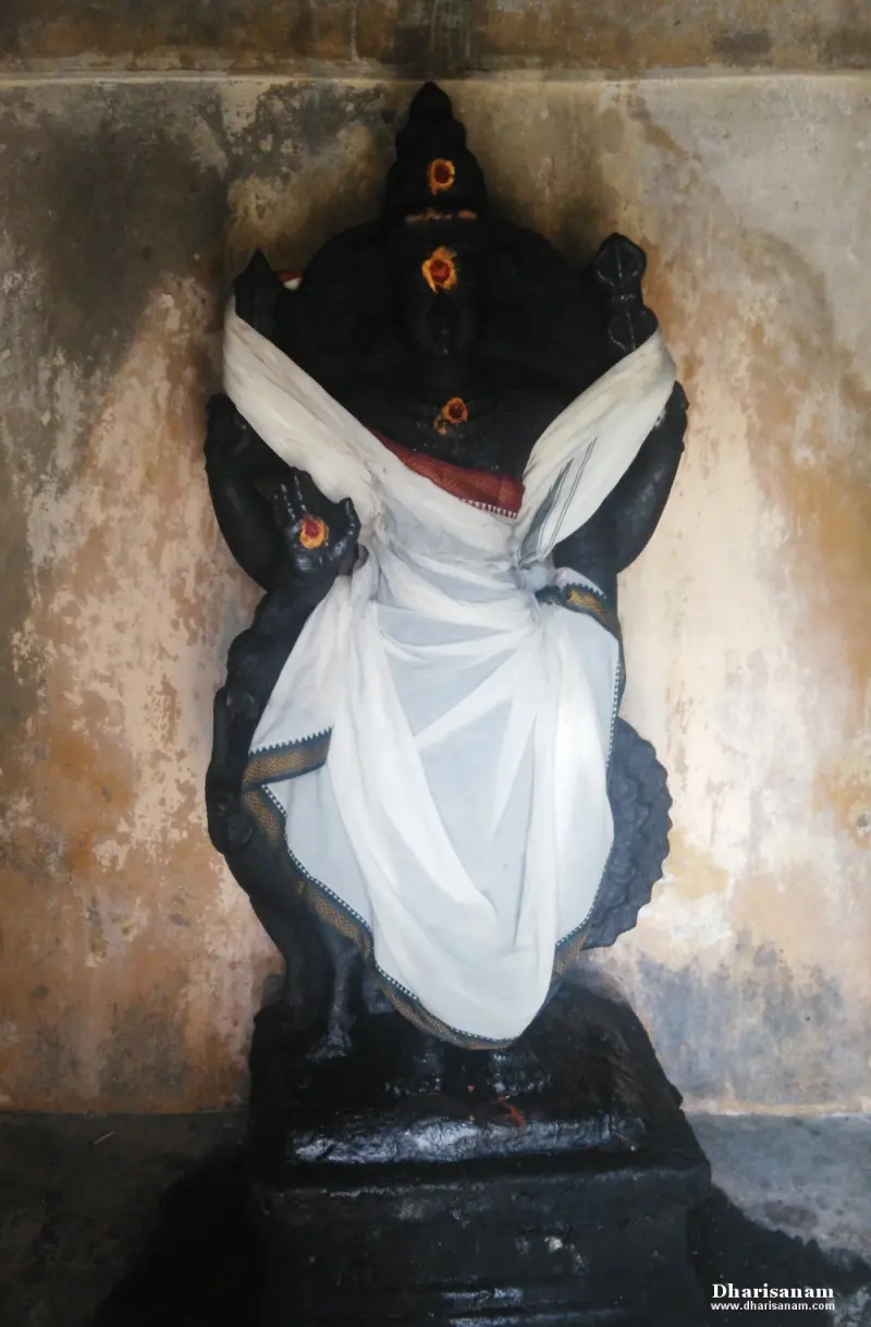 A tall four-armed dark idol draped in white cloth, standing against a wall.