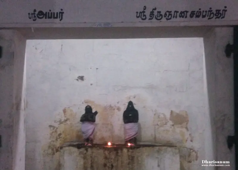 Two small dark images in cloth stand on a ledge with two lit lamps, under their Tamil names.