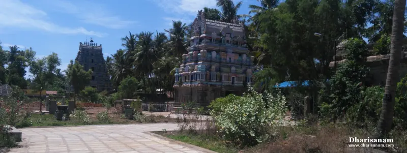 A coloured gateway tower among palm trees with a dark stone tower further behind, seen from a paved path