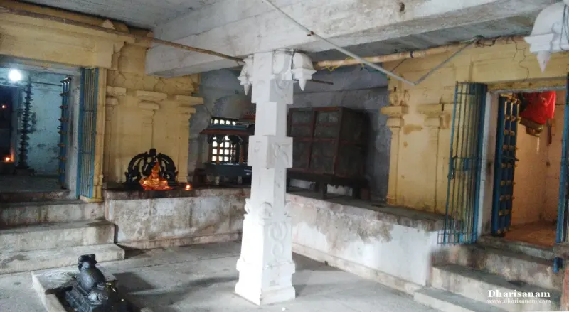 A covered stone corridor with a white pillar, a small Nandi at lower left, a lit lamp and shrine doorways on both sides.