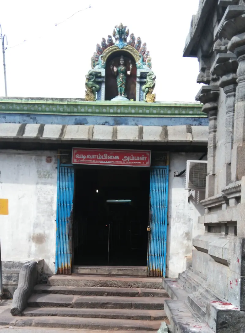 A shrine doorway with blue shutters and a red name board reading Vadivambikai Amman, with a goddess figure above.