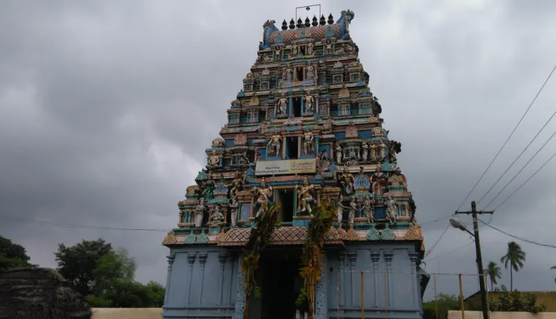 A tiered gopuram covered in painted sculptures, seen under a cloudy sky with coconut palms beside it.