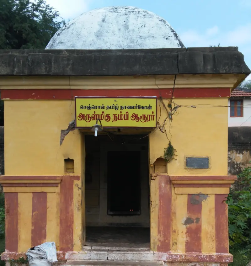 A small yellow shrine with red stripes and a domed top; a Tamil name board hangs above the open doorway.