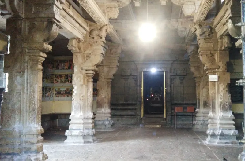 A stone hall with carved pillars; paintings line the left wall and a lit doorway leads to the sanctum at the far end.