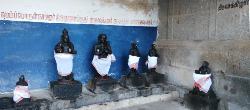 Row of small dark stone figures in white cloths before a blue wall with name boards