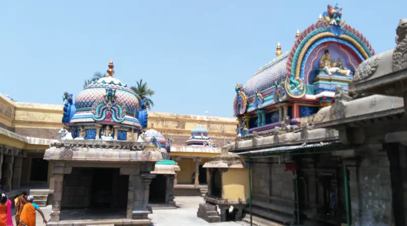 A paved courtyard with a painted dome on the left, an arched shrine roof on the right, and visitors walking