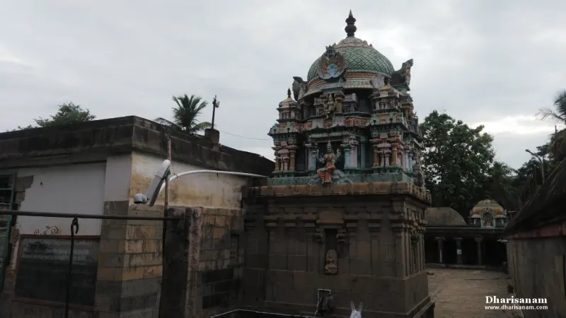 A shrine tower with a green dome and small figures, beside a low plastered wall under a grey sky.