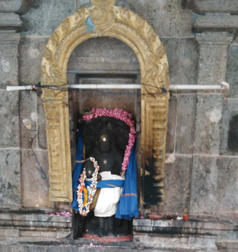 A dark idol stands in a stone niche under a gilded arch, with flower garlands and a blue and white cloth.