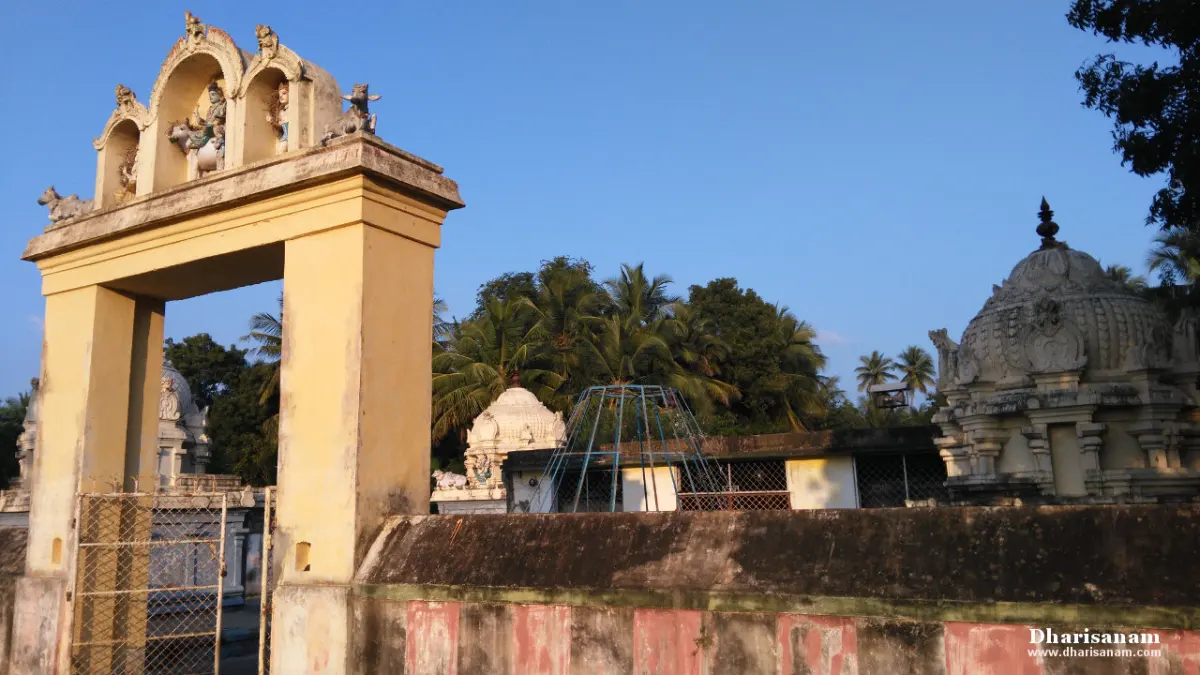 A yellow gateway arch with small figures on top, a boundary wall and shrine domes behind it under a blue sky