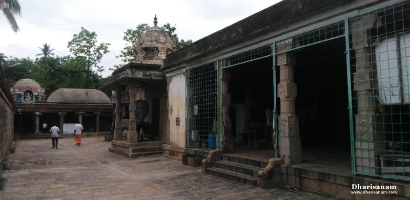 A paved courtyard with a pillared hall behind green grilles and a small shrine tower; a visitor walks at left.