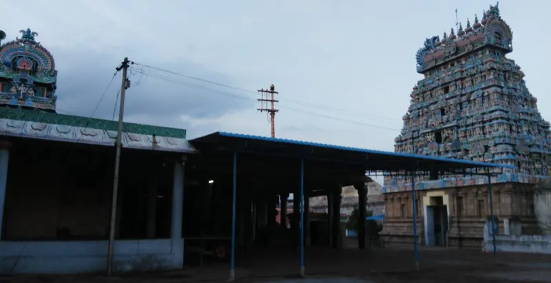 A flagstaff stands in an open courtyard beside a tin-roofed shed; a tall sculpted tower rises at right.