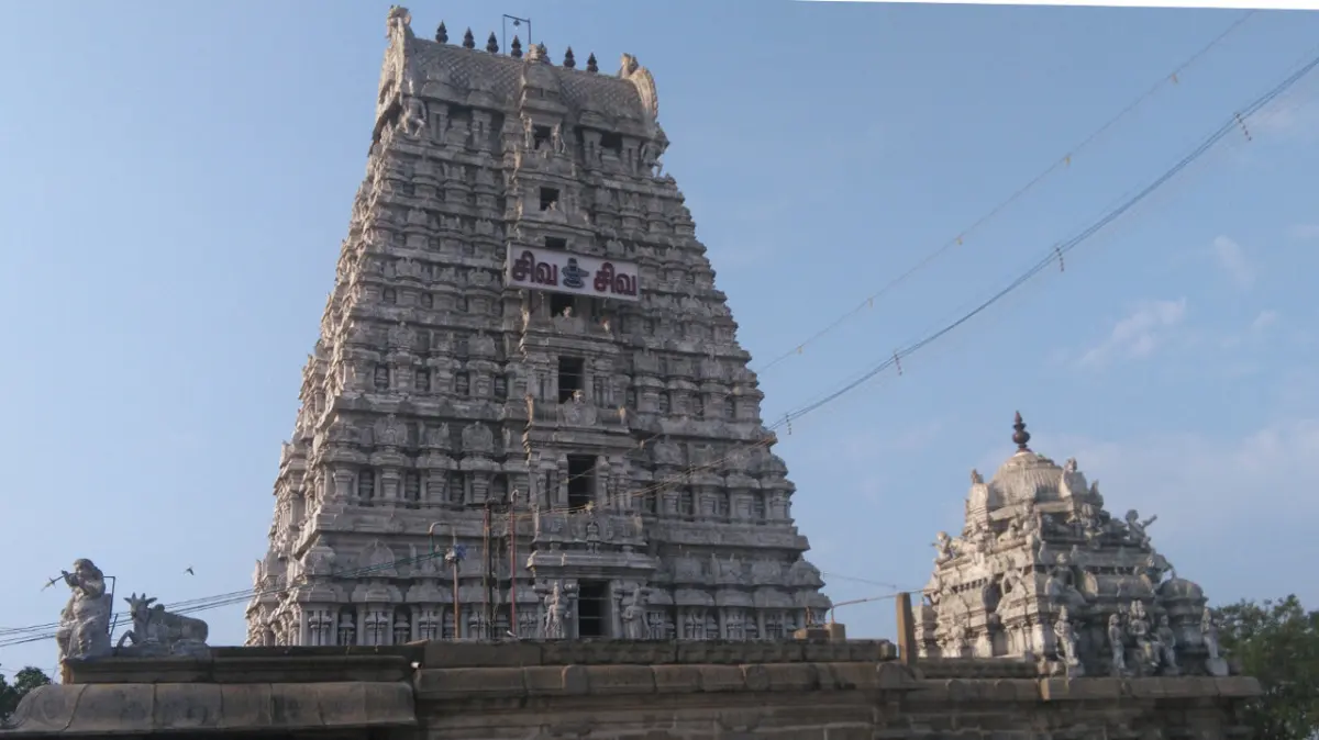 A tall tiered gopuram of grey stone with a Tamil sign, and a smaller domed tower to its right