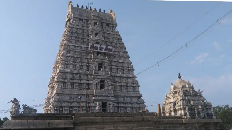 A tall tiered gopuram of grey stone with a Tamil sign, and a smaller domed tower to its right