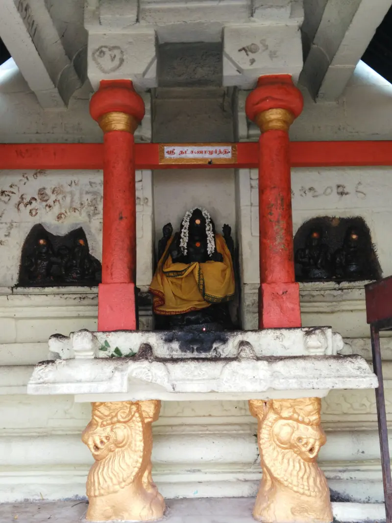 A dark idol in yellow cloth and a flower garland between red pillars, with a name board above.