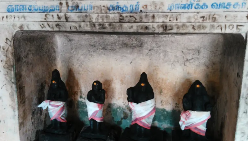 Four small black stone figures with white and pink cloths stand in a wall niche under a Tamil inscription.