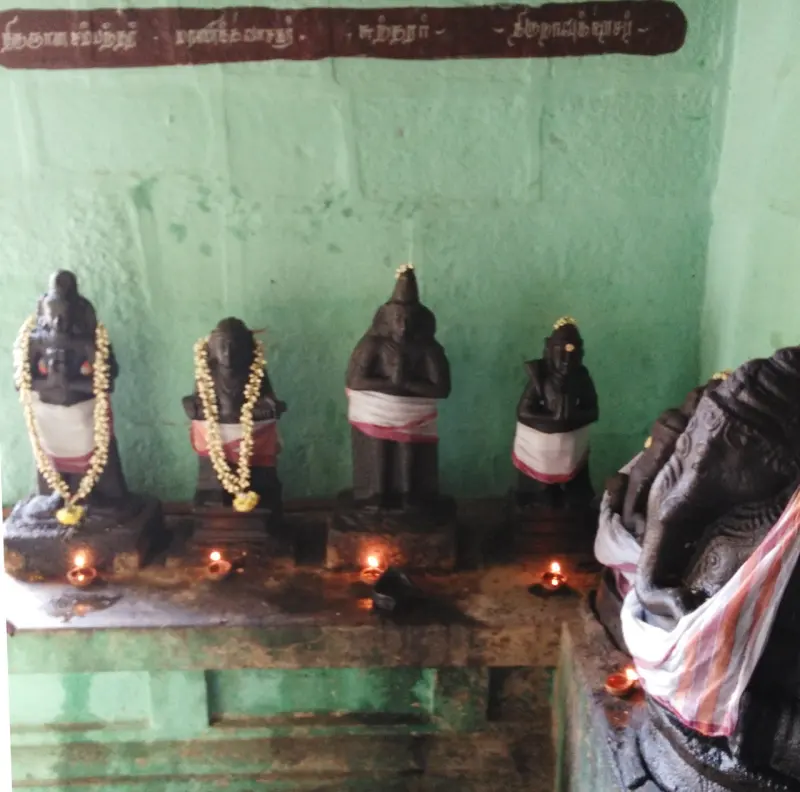 Four small dark idols with jasmine garlands and lamps below a Tamil name board, with a Vinayakar's trunk at right.