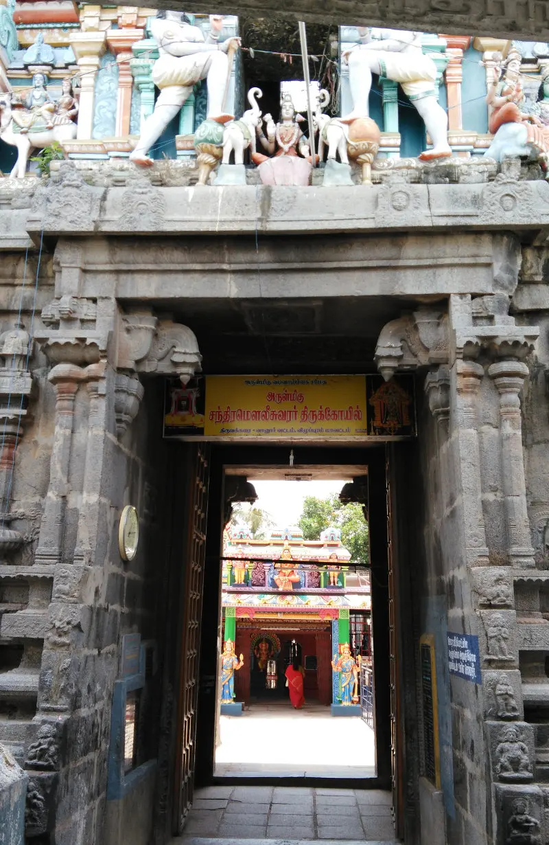 A stone gateway with figures on top and a Tamil name board naming the temple, opening to a colourful shrine beyond.
