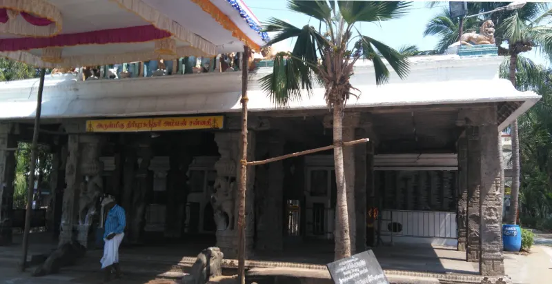 A stone pillared hall with a yellow Tamil name board and a palm tree propped with a pole.