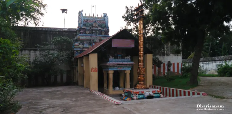 A tall gold-coloured pillar beside a small roofed mandapam, with a coloured tower rising behind the wall