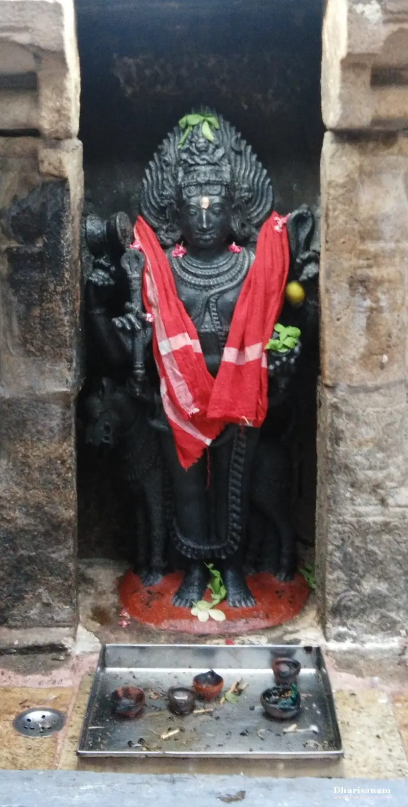 A black stone idol in a niche, with a red cloth over the shoulders and a tray of small lamps below