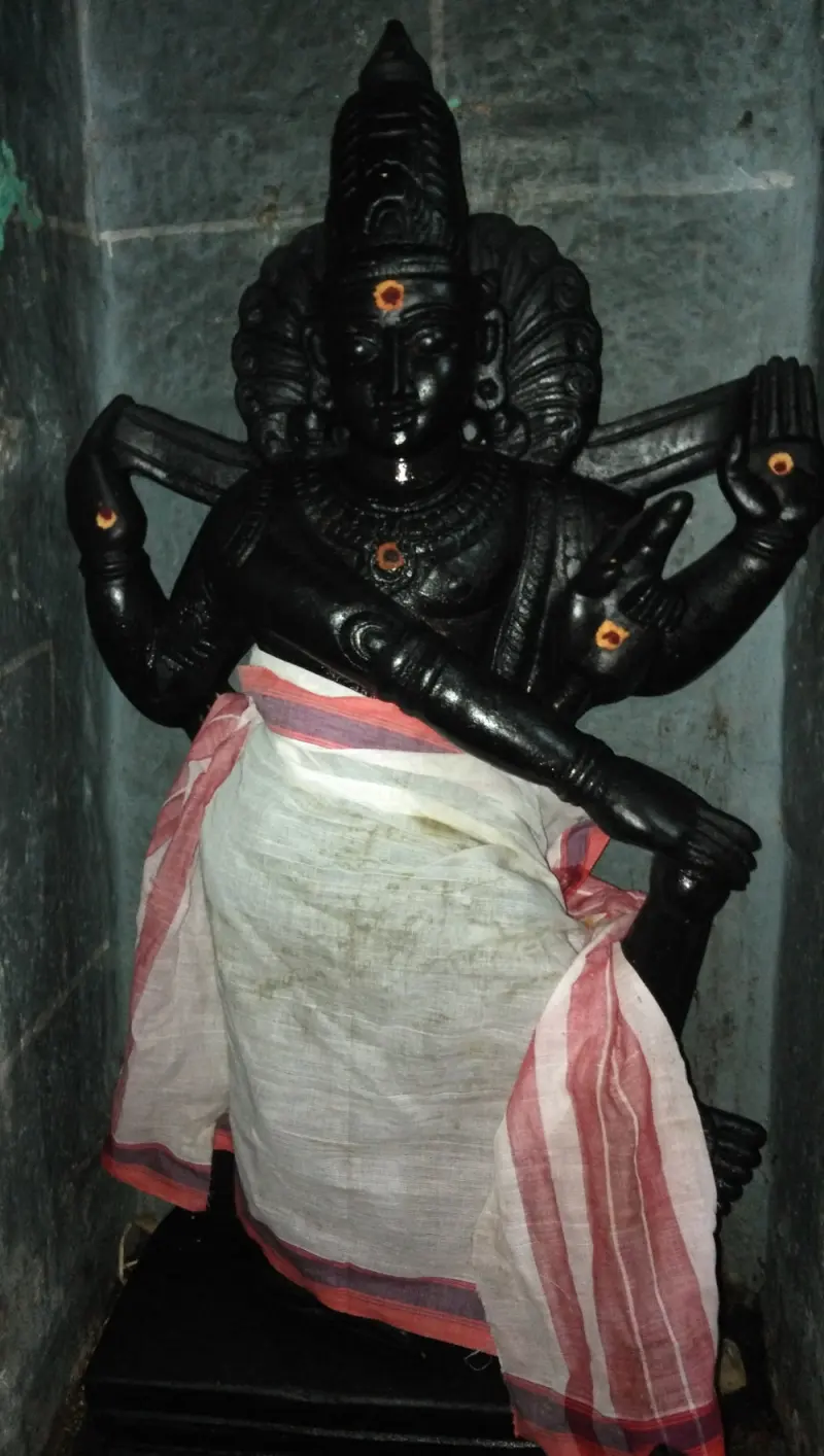 A four-armed black stone idol with a tall crown, wearing a white cloth with a red border, standing in a niche.