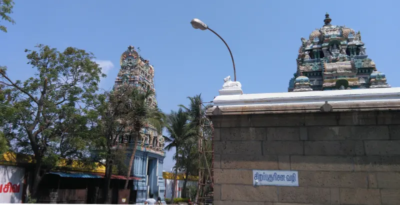 A stone wall with a Tamil sign, a small Nandi on top, and two towers against a blue sky.