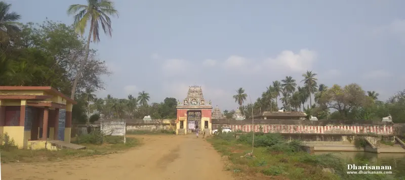 A dirt road leading to the temple's red and yellow gateway, with palm trees and a boundary wall beside it