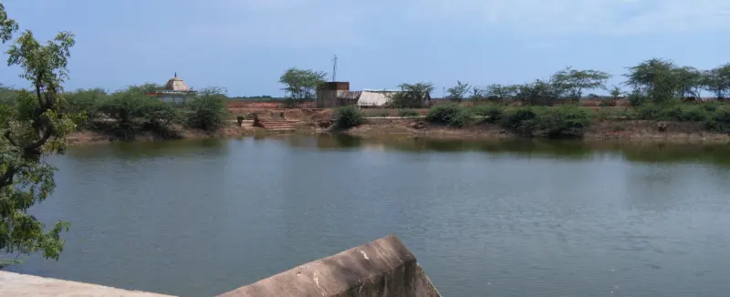 A wide still tank with steps on the far bank and a small shrine among trees
