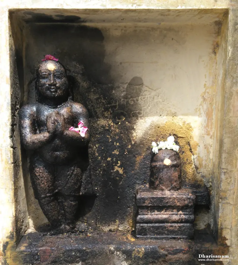 Stone figure with folded hands, wearing a flower, stands in a niche beside a small lingam